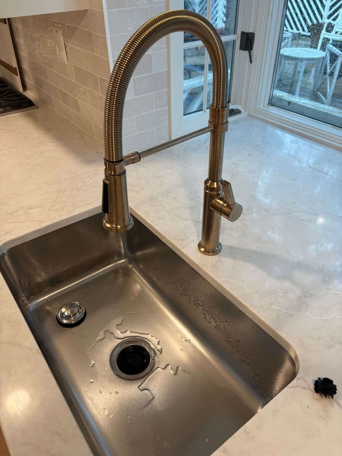 Stainless steel kitchen sink with gold faucet against a light countertop and tiled backsplash, near a window.