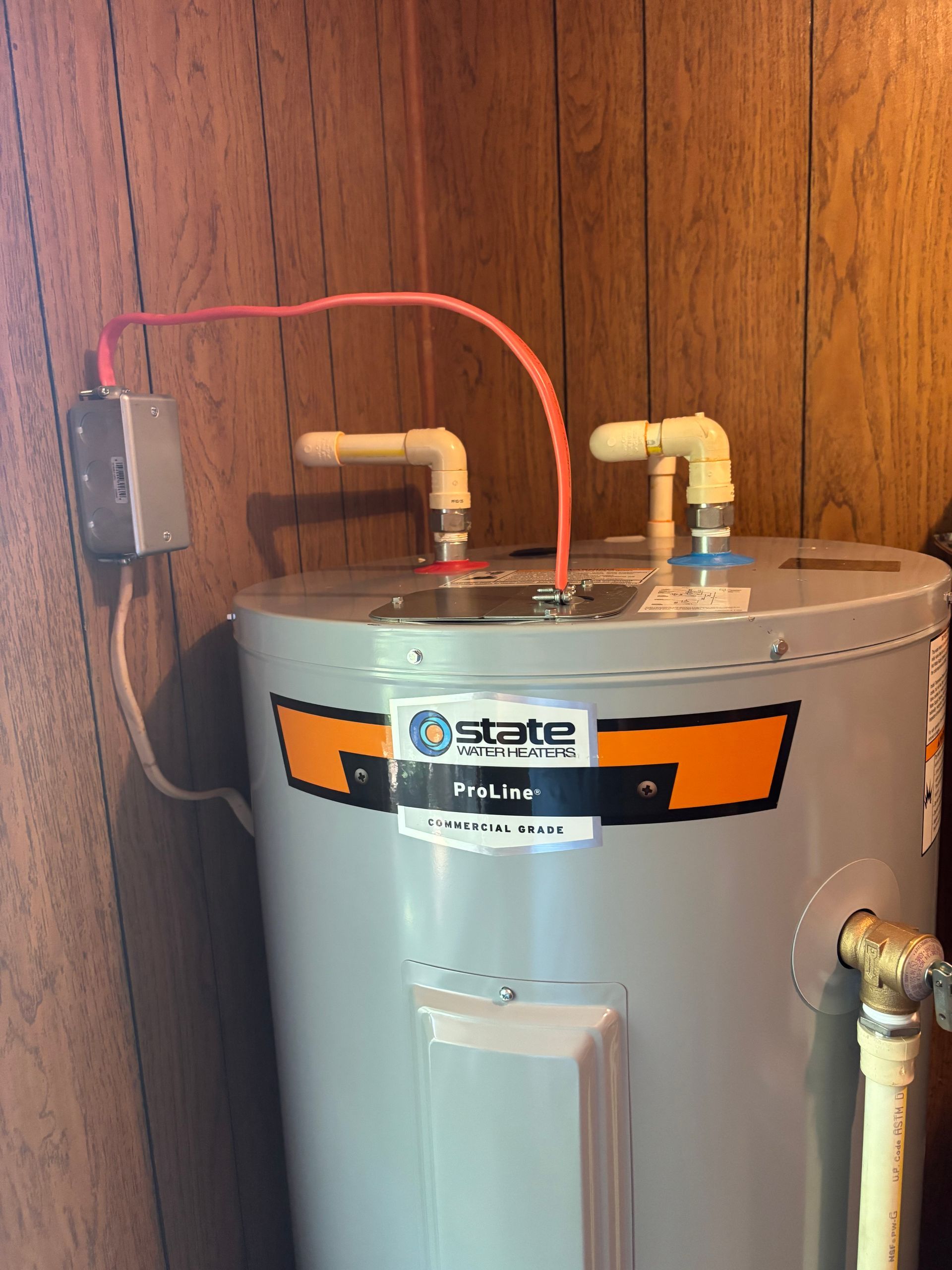 Water heater with pipes, electrical box, and red wire against wood paneling.