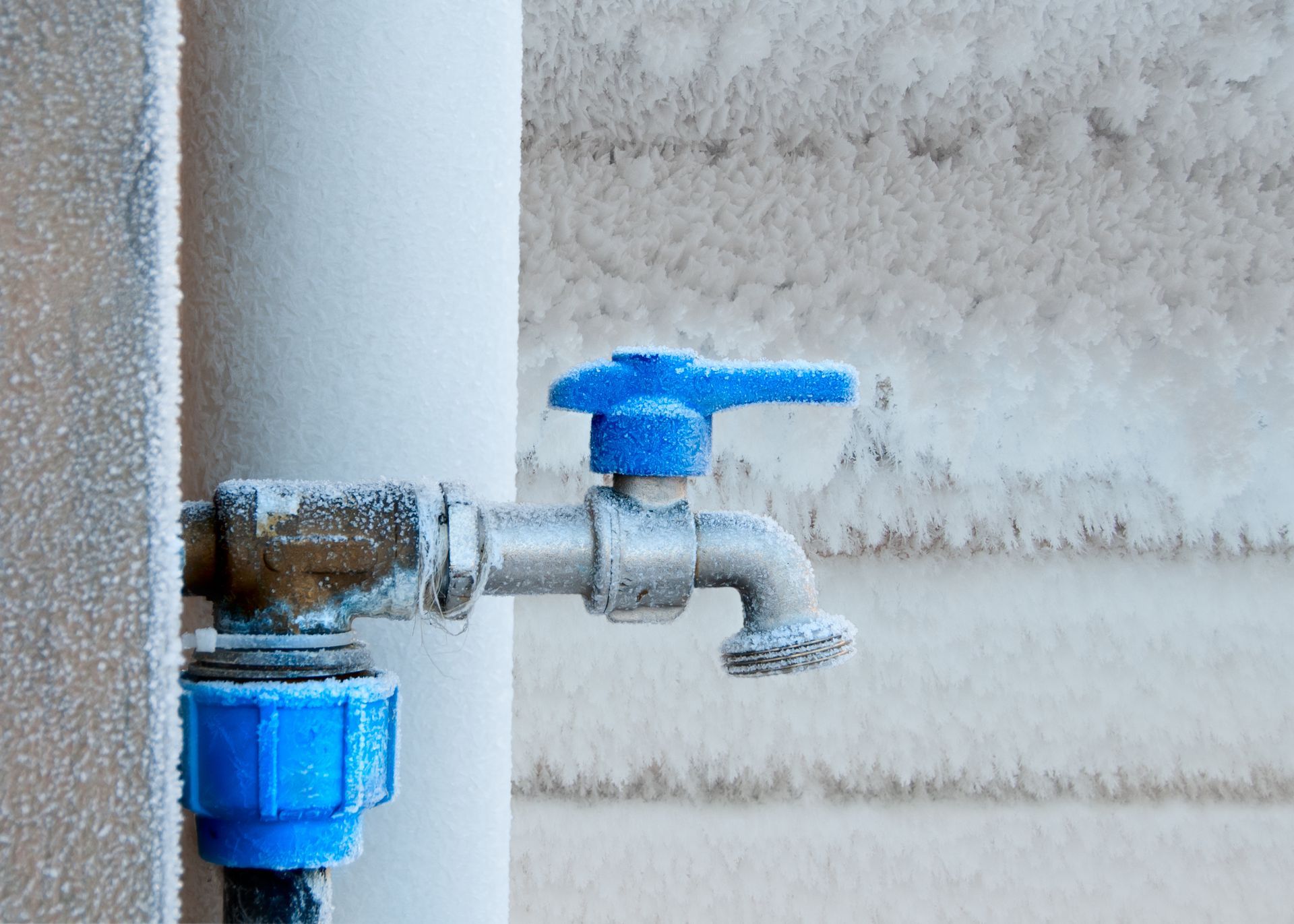 Frozen outdoor water tap with blue handle; snow-covered pipes and wall.