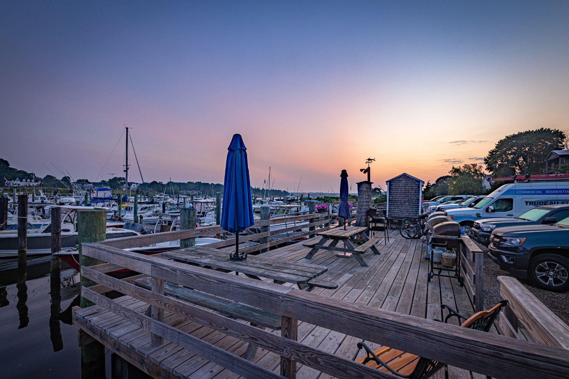 A wooden deck with tables and umbrellas in a marina at sunset.