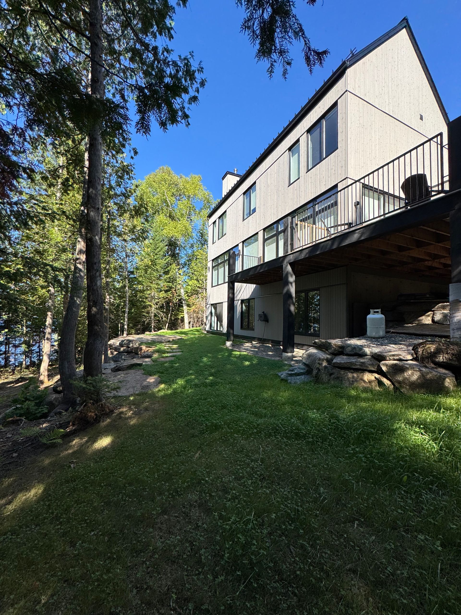 Maison à deux étages avec un revêtement extérieur clair, une terrasse noire et une pelouse verdoyante par une journée ensoleillée.