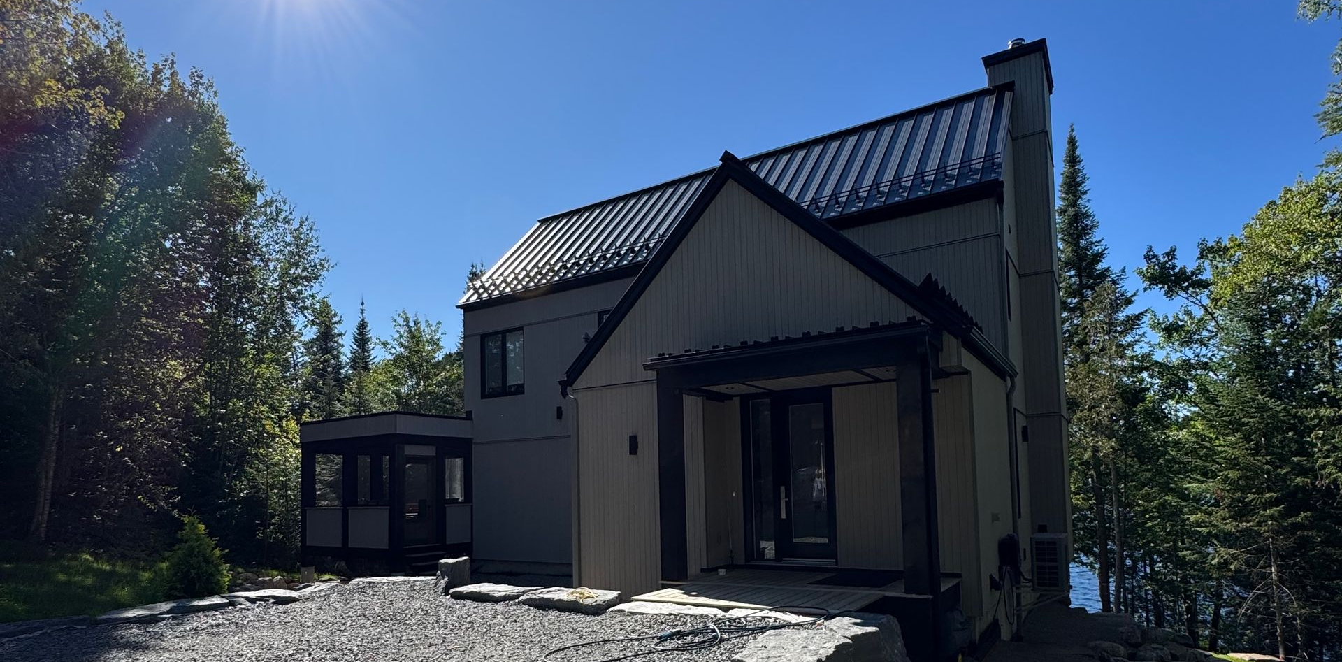 Maison moderne à deux étages avec toit noir, cheminée et entrée sous un ciel bleu clair, entourée d'arbres.