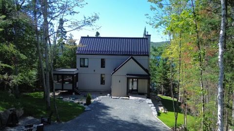 Maison à deux étages avec toit foncé et bardage gris clair, nichée dans les arbres et dotée d'une allée de gravier.