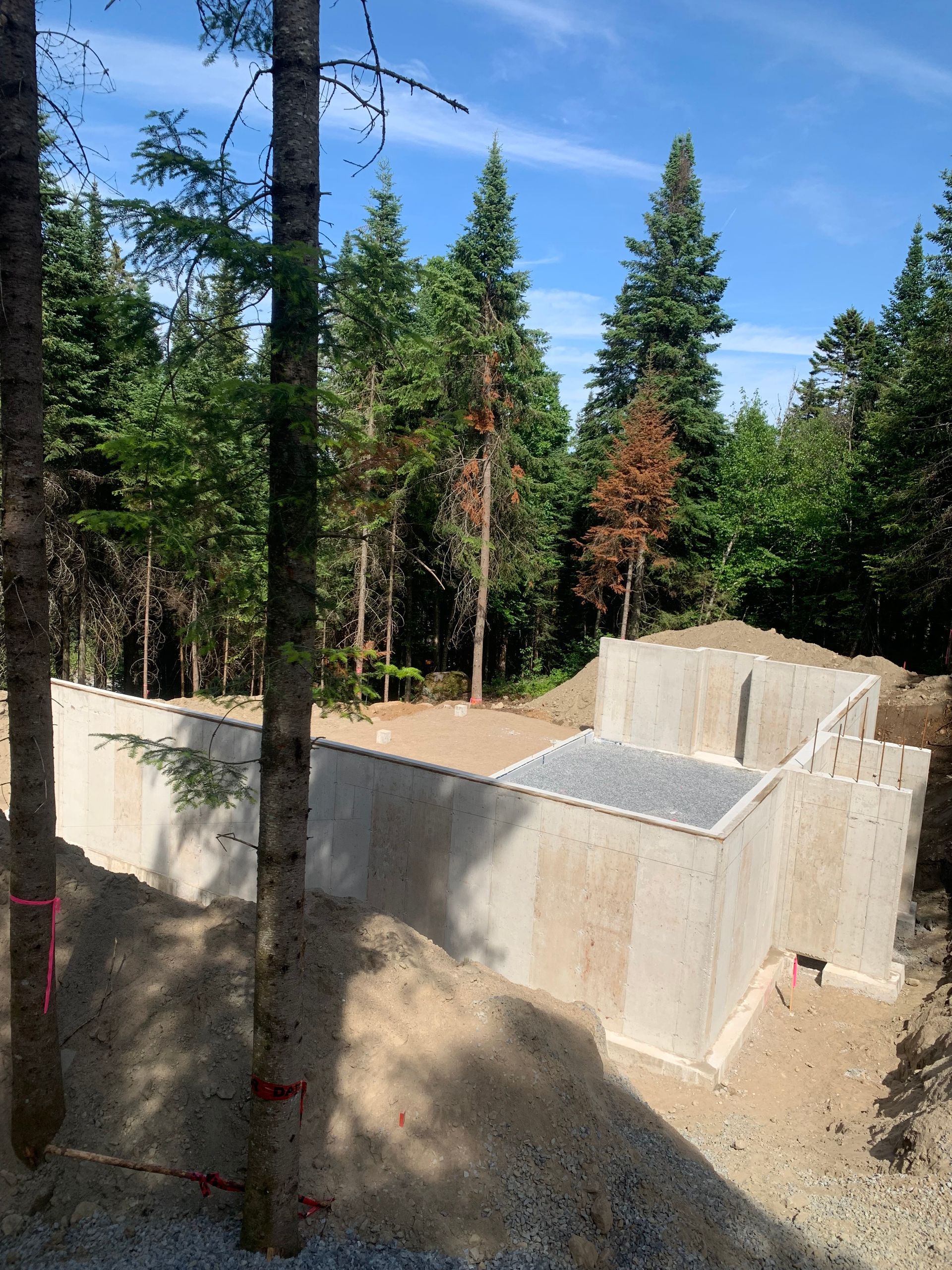 Construction de fondations en béton dans une zone boisée, avec des arbres au premier plan et à l'arrière-plan, par une journée ensoleillée.