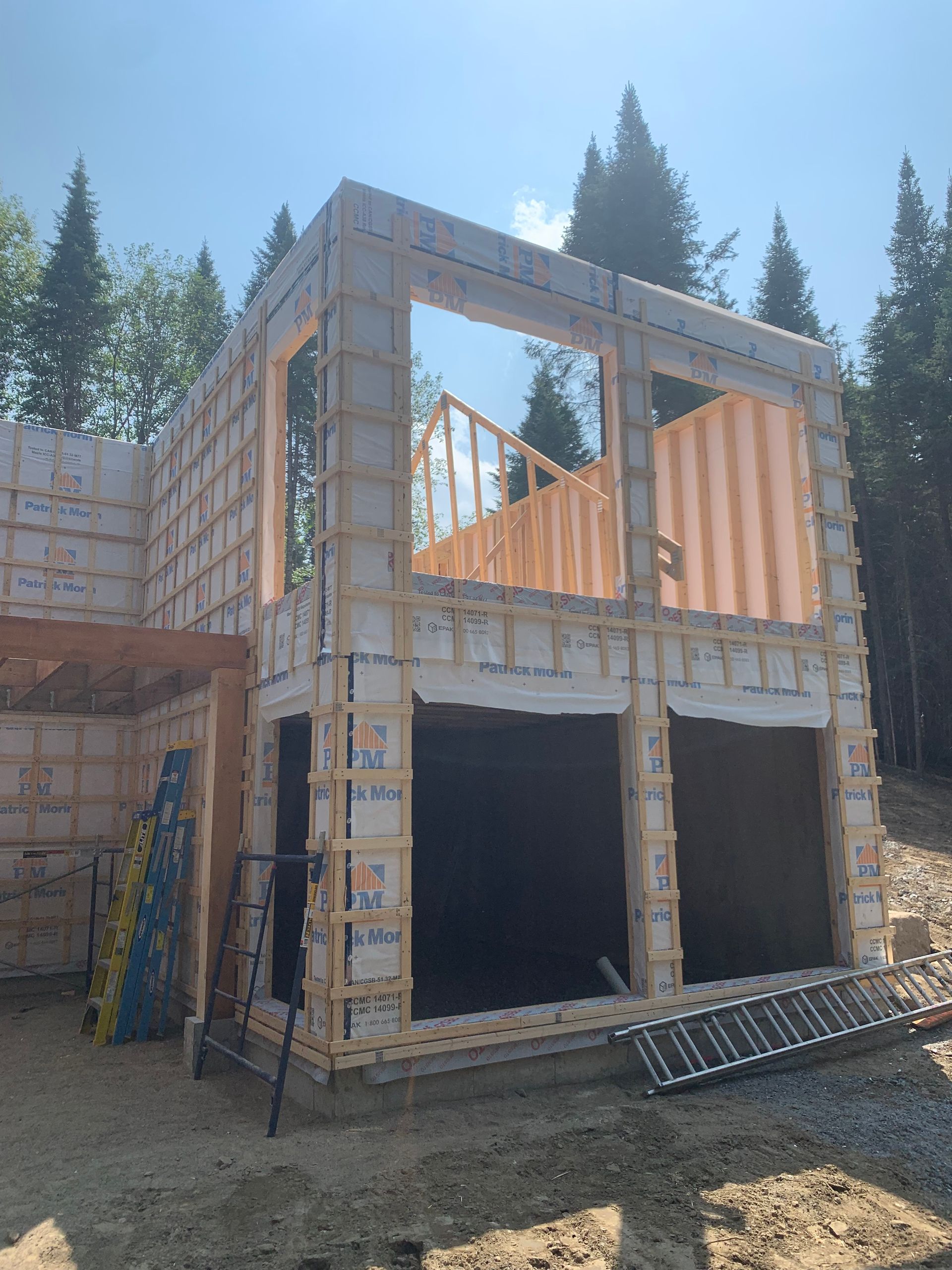 Bâtiment de deux étages en construction, avec une charpente en bois apparente et de grandes ouvertures vitrées, sur fond de forêt.
