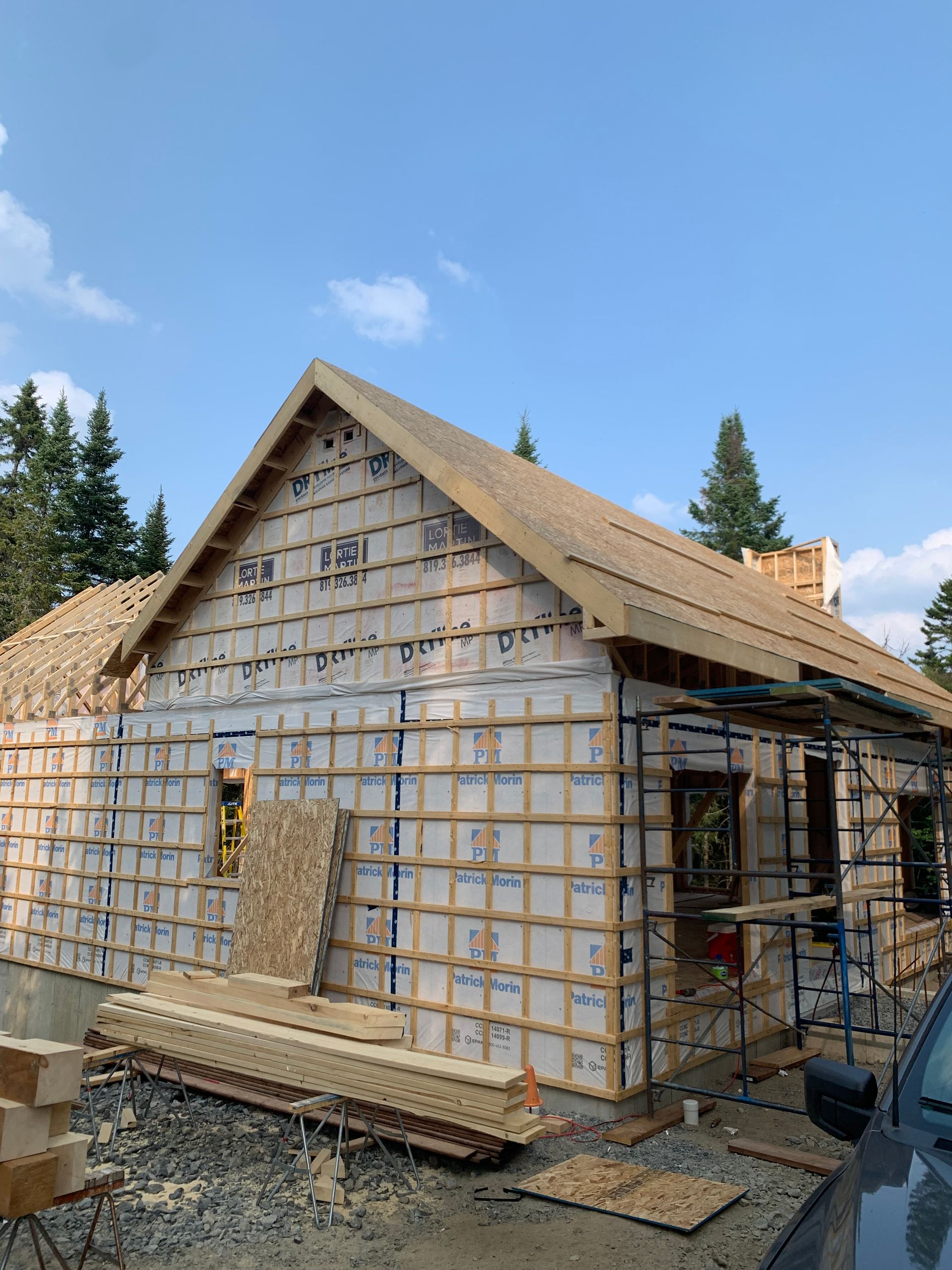Chantier : Extérieur d'une maison avec isolation et charpente de toit sur fond de ciel bleu, entourée d'arbres.