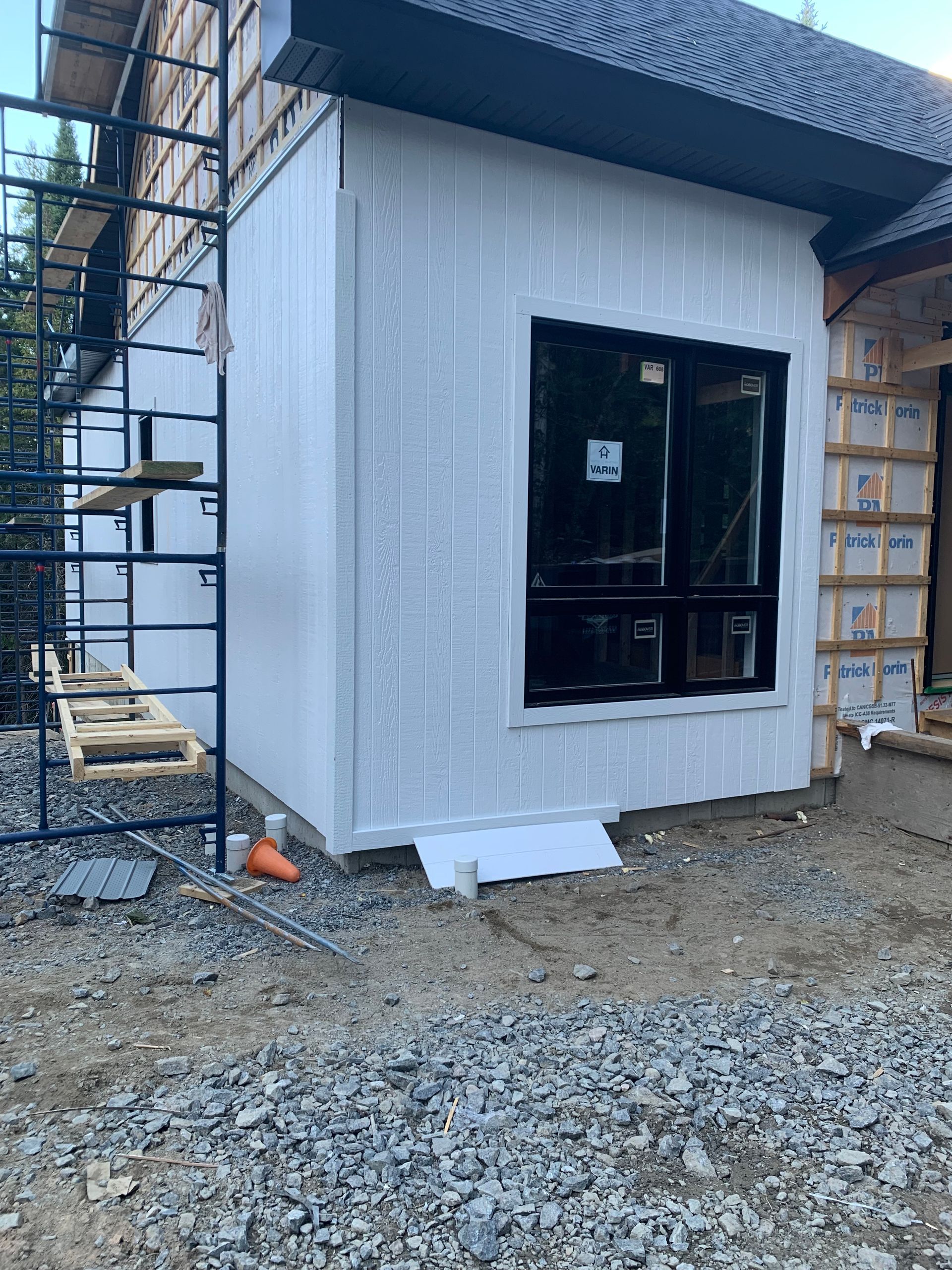 Extérieur d'un bâtiment en construction, avec un bardage blanc, un encadrement de fenêtre noir et des échafaudages.