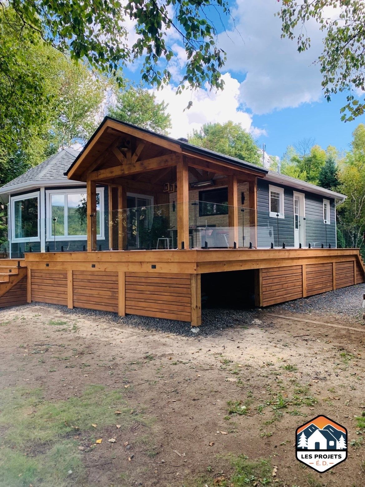 Terrasse en bois avec un espace couvert prolongeant la maison, dotée d'une rambarde en verre et de panneaux décoratifs, sous un ciel bleu.
