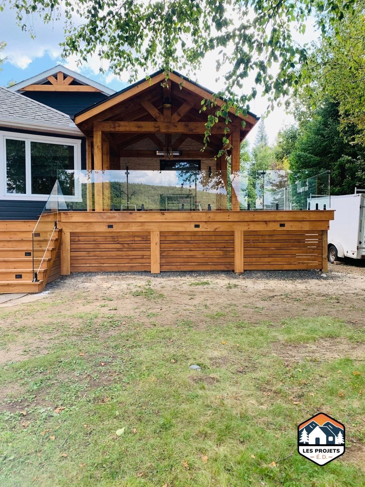 Terrasse en bois avec rambarde en verre et zone couverte, attenante à une maison à façade bleue, à l'extérieur.