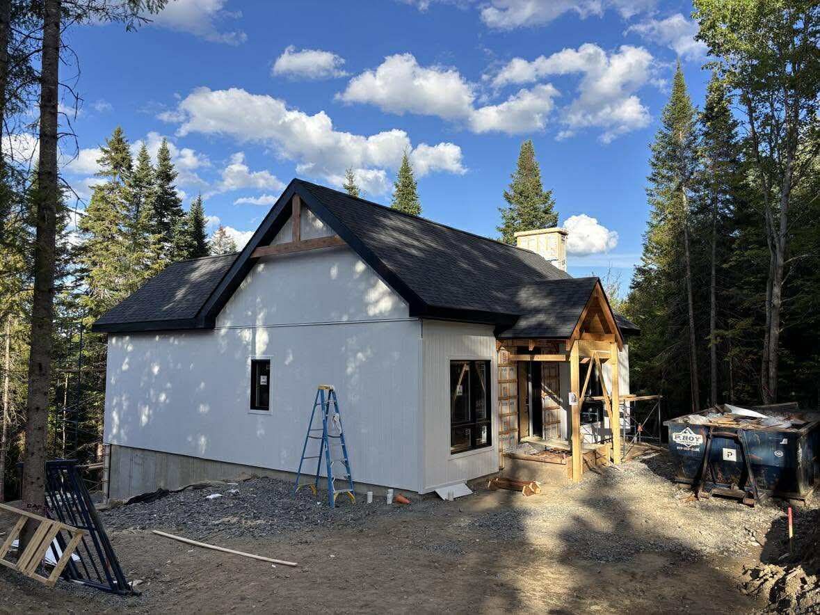 Maison en construction dans une zone boisée, avec un bardage blanc, un toit foncé et des échafaudages autour de l'entrée.