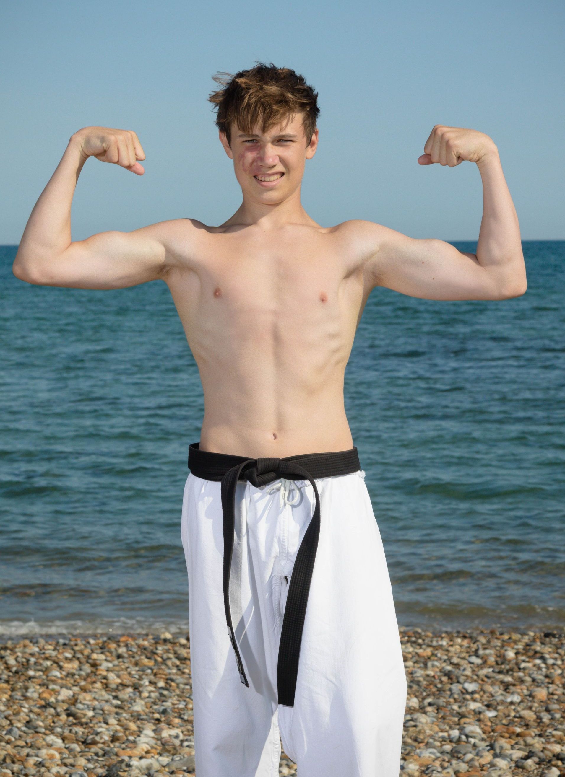 A shirtless young man is flexing his muscles on the beach.