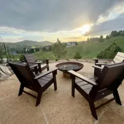 Four dark Adirondack chairs surround a fire pit on a stone patio overlooking a hilly landscape at sunset.