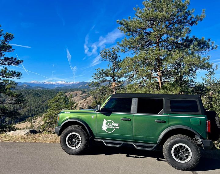 Green Ford Bronco parked on a scenic mountain overlook with a backdrop of trees, mountains, and blue sky.