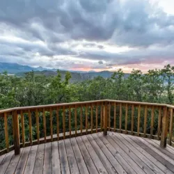 Wooden deck overlooking a forest and mountains under a cloudy sunset sky.