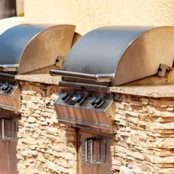 Two stainless steel outdoor grills built into a stone wall with tan trim.