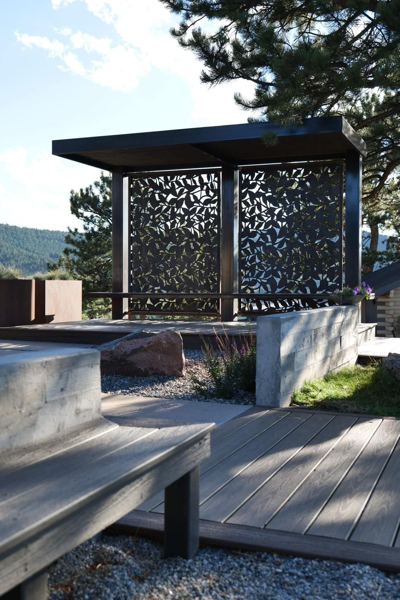 Outdoor pavilion with patterned screens, concrete bench, and wooden walkway against a hillside.
