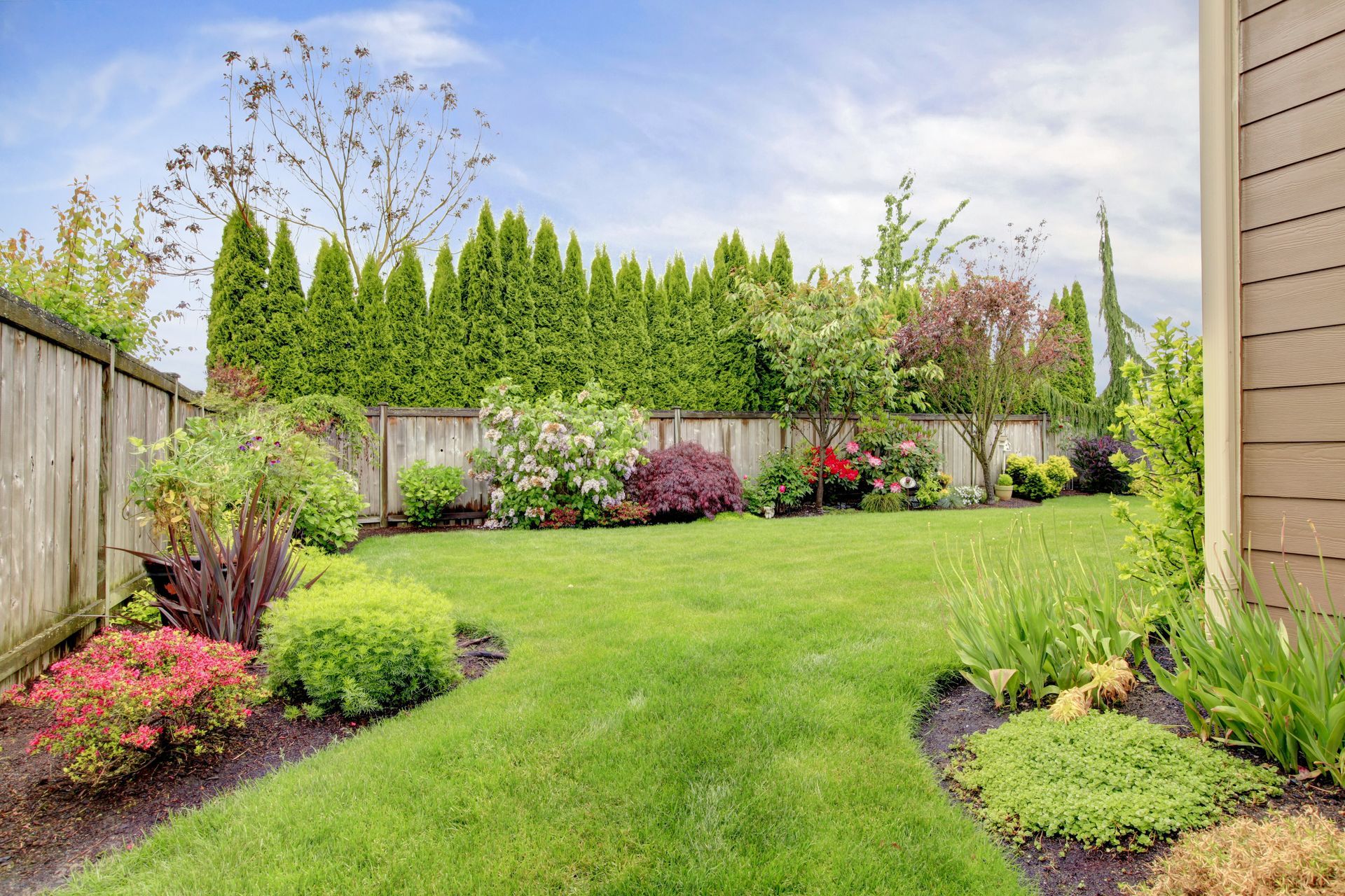 Green lawn, garden beds with various plants, wooden fence, and tall evergreens under a partly cloudy sky.