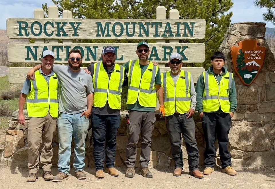 Six people in safety vests pose in front of the Rocky Mountain National Park sign.