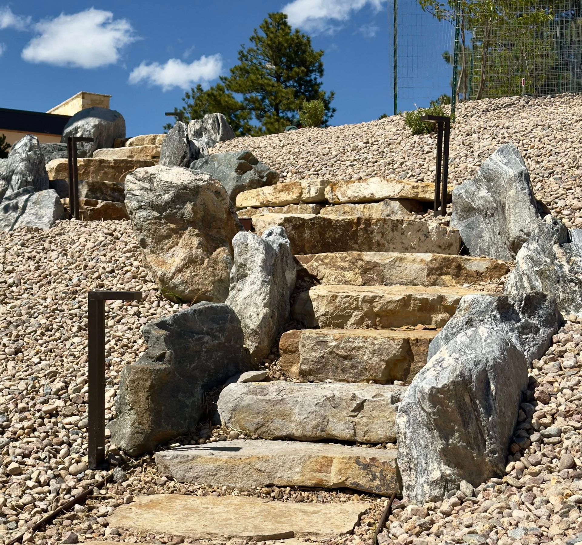 Stone steps ascend a gravel-covered hillside, flanked by large boulders and small, brown path lights.