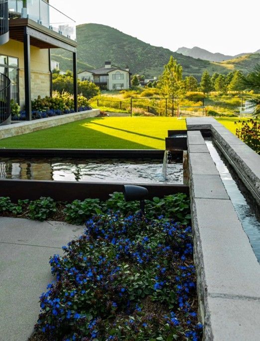Lush green lawn and water features with a mountain backdrop, complemented by a modern home and blue flowers.