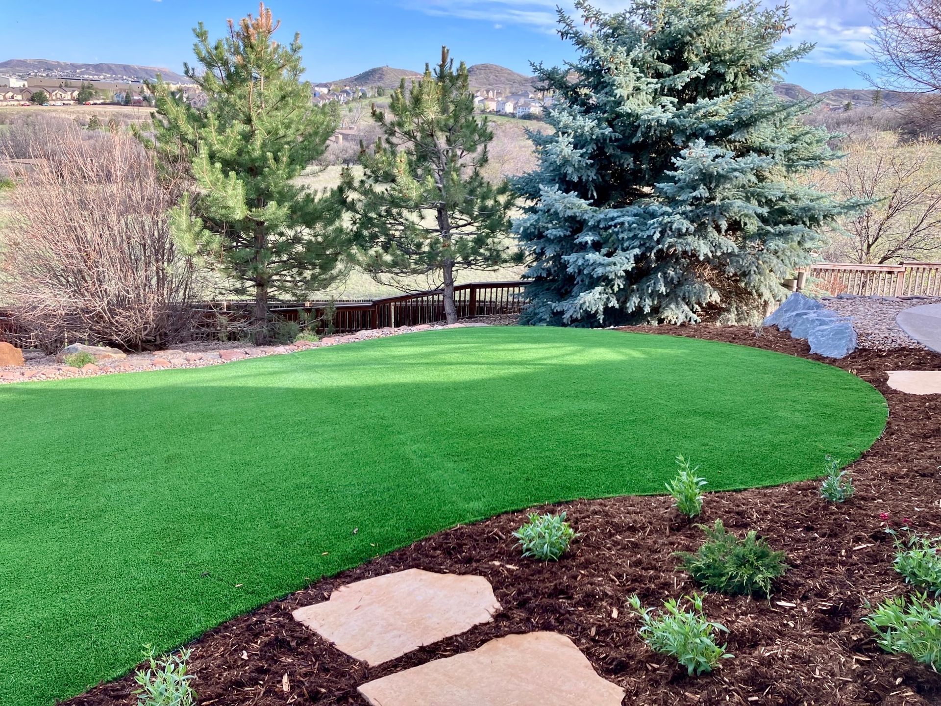 Green artificial turf lawn with mulch and stepping stones, trees and mountain backdrop.