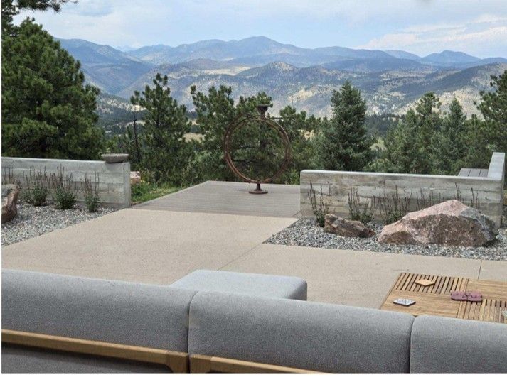 Patio overlooking mountains; gray concrete, trees, a sofa, and large rock are in view.