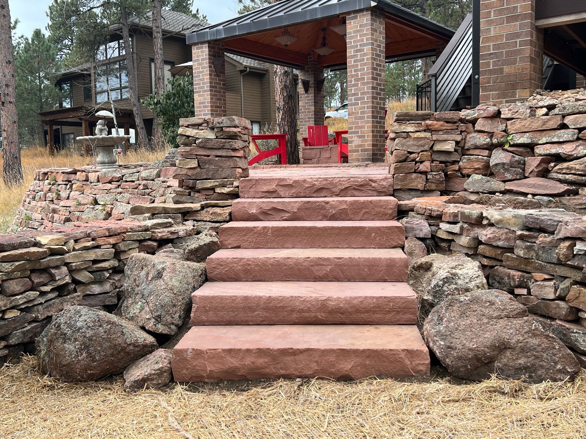 Stone steps leading up to a brick and wood structure with red chairs, surrounded by stone walls and a house.