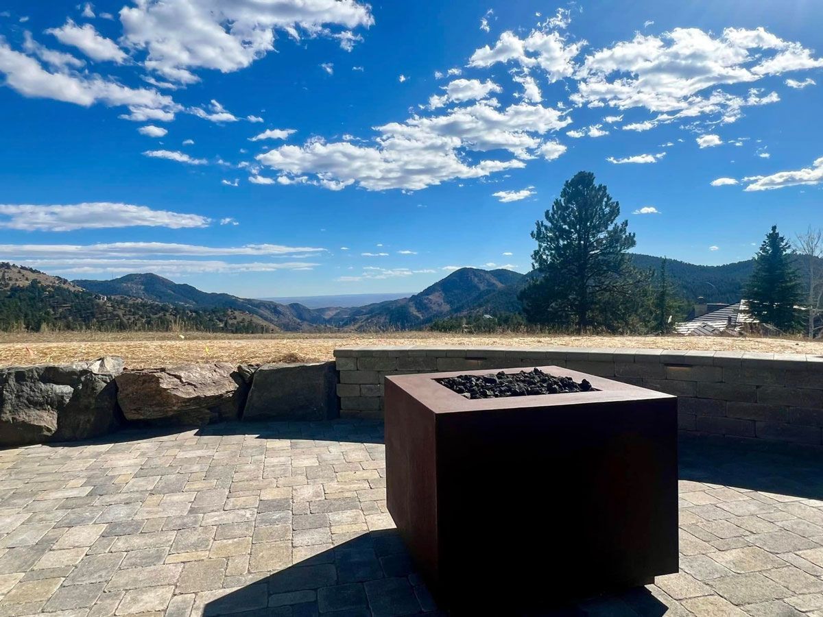Patio with a modern fire pit, stone patio, and mountain view under a blue, cloudy sky.