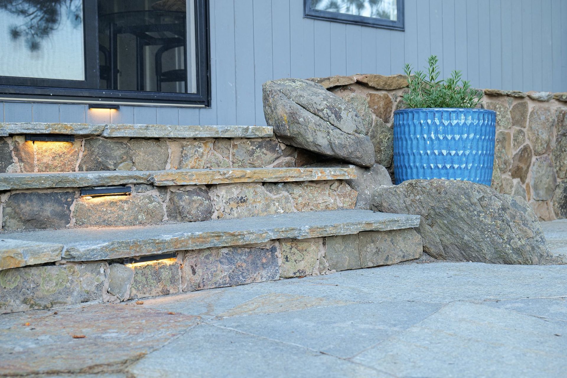 Stone steps with built-in lighting leading to a blue planter. Grey siding and dark window in background.