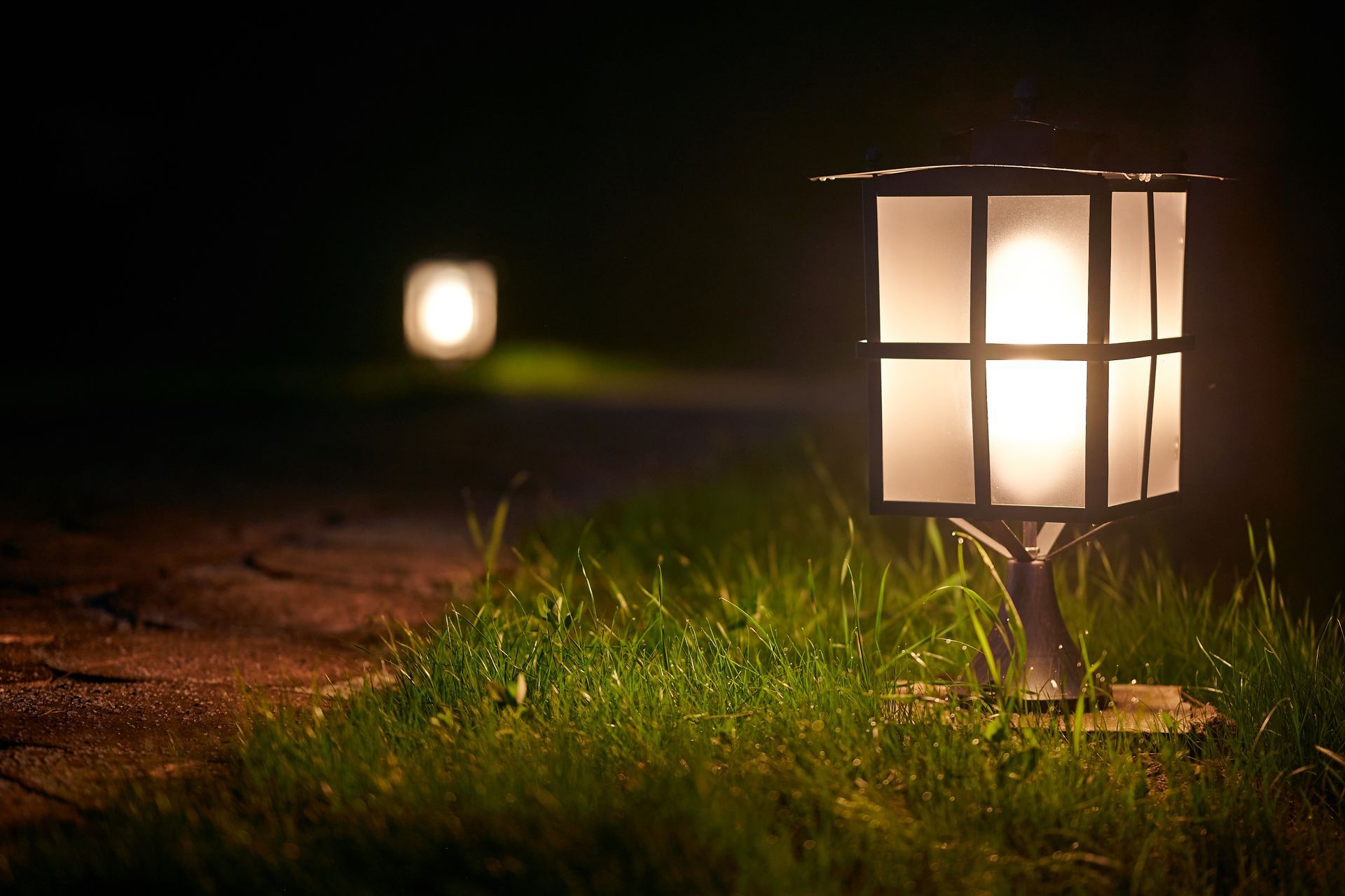 Lawn lamp glowing at night; another lamp glows dimly in the distance along a path.
