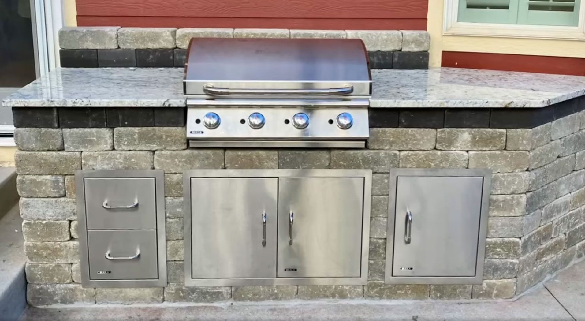 Outdoor kitchen with stainless steel grill, doors, and drawers built into a brick structure.