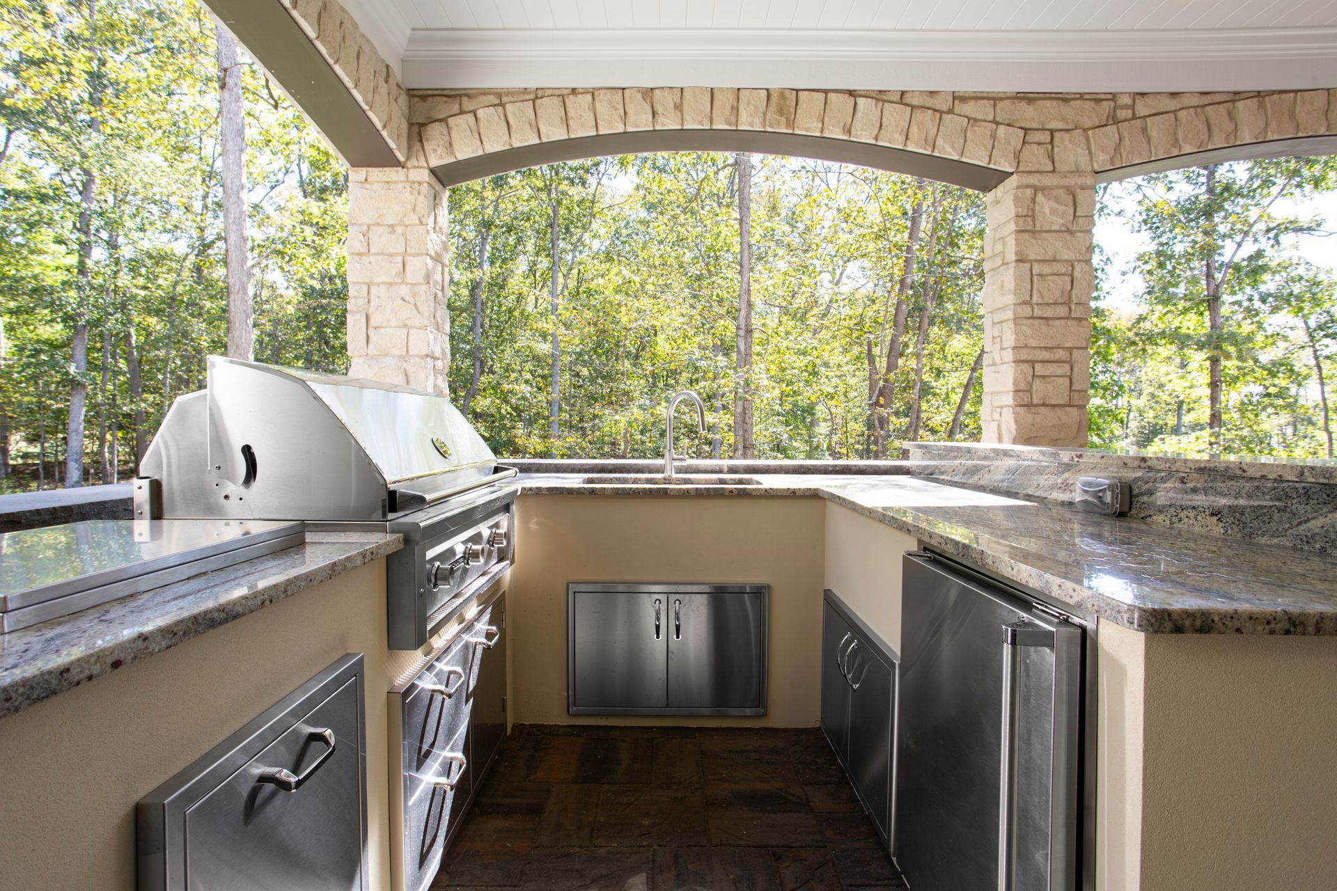 Outdoor kitchen with built-in grill, countertops, and stainless steel appliances, open to a wooded view.