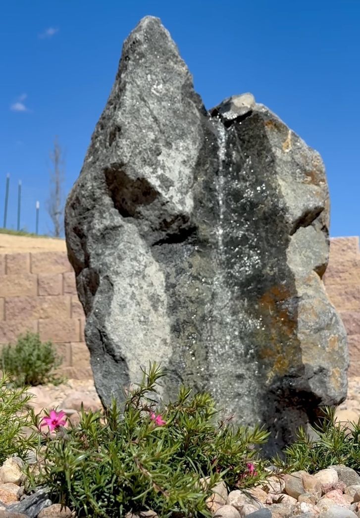Large, gray rock fountain with water cascading down, surrounded by flowers and a blue sky.