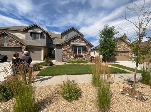 House exterior with landscaped yard, two people standing in front of it. Sunny, blue sky.