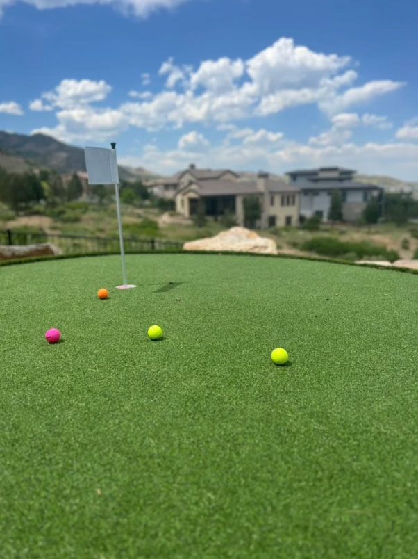 Green putting green with several colorful golf balls and a flag. Houses and hills in the background on a sunny day.