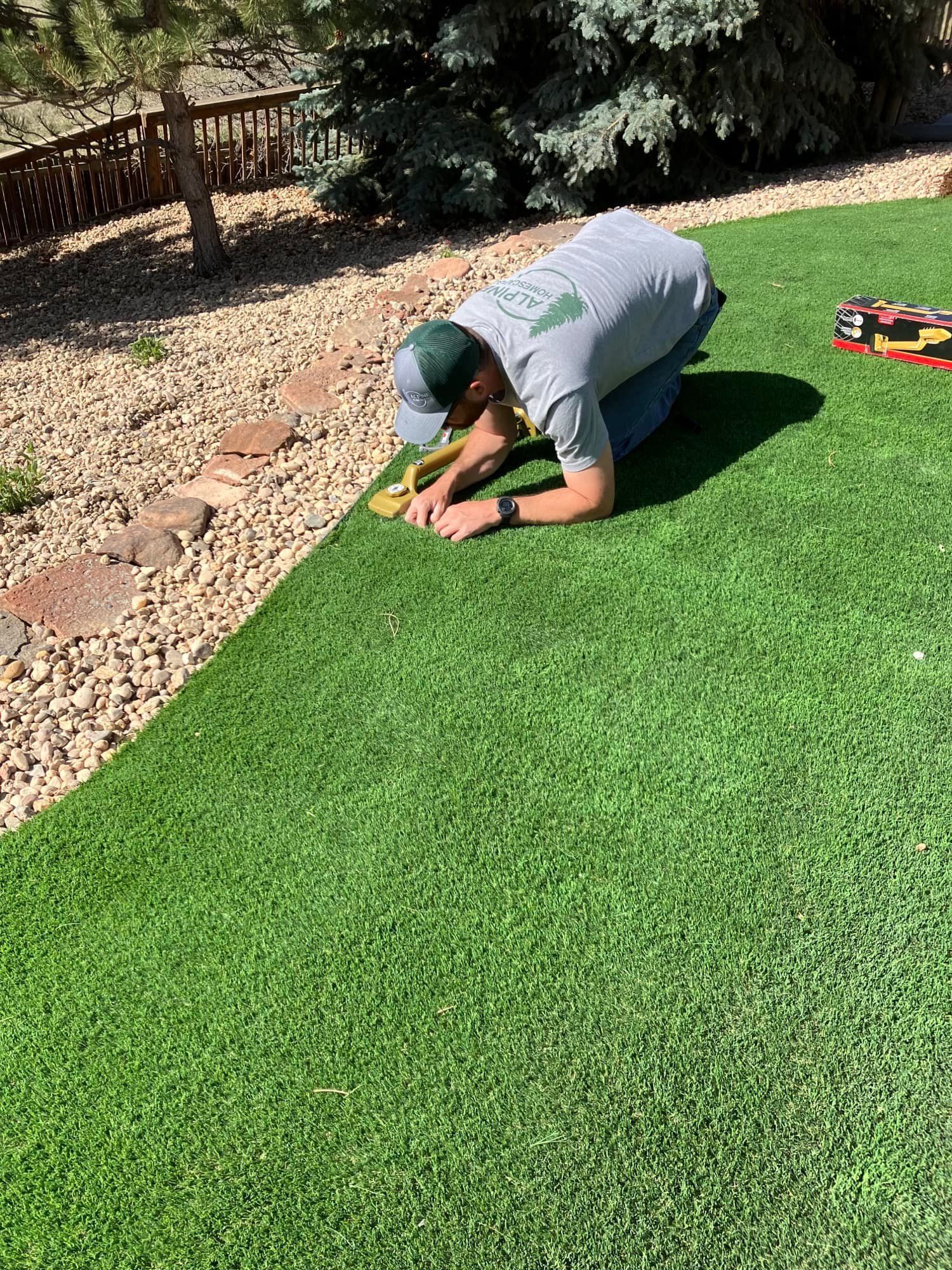 Person kneeling, trimming artificial turf edge with a knife in a backyard.