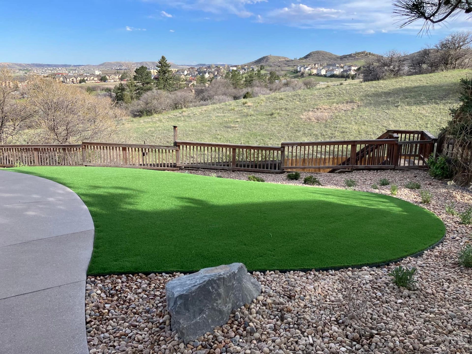 Green artificial turf yard with wood deck overlooking a hillside and houses under a blue sky.