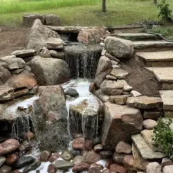 Waterfall cascading over rocks into a pool filled with stones. Stone steps ascend to the right, green grass in background.