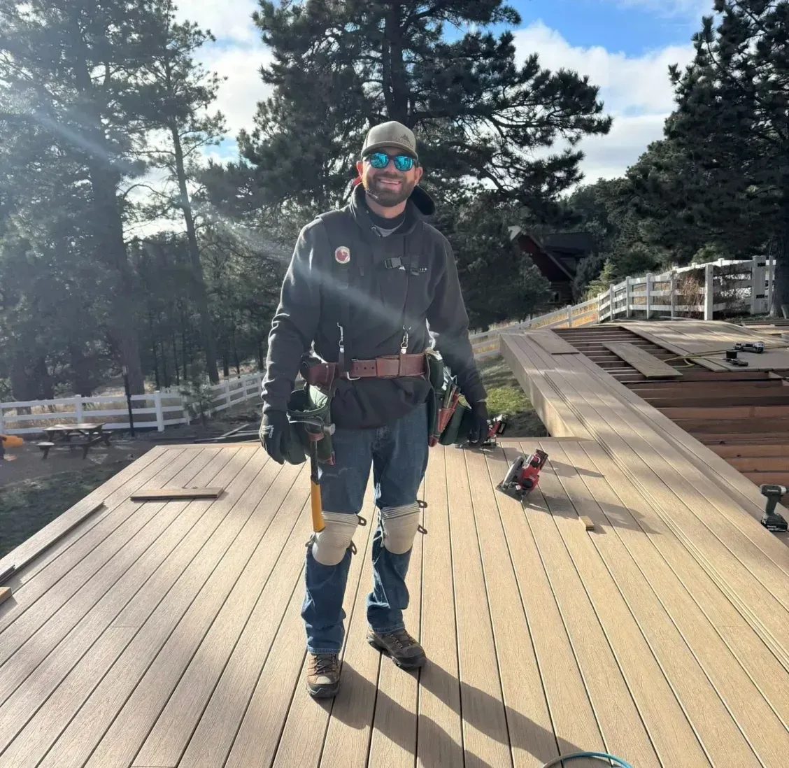 Roofer stands on a partially-built wooden deck, wearing work gear. Blue sky and trees in the background.