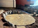 Stone patio with rock border surrounded by dirt, near a building.
