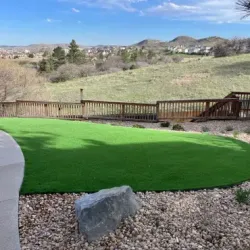 Green artificial turf yard with wood deck overlooking a hillside and houses under a blue sky.