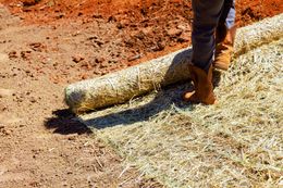 A person rolls out erosion control matting on a red dirt slope.