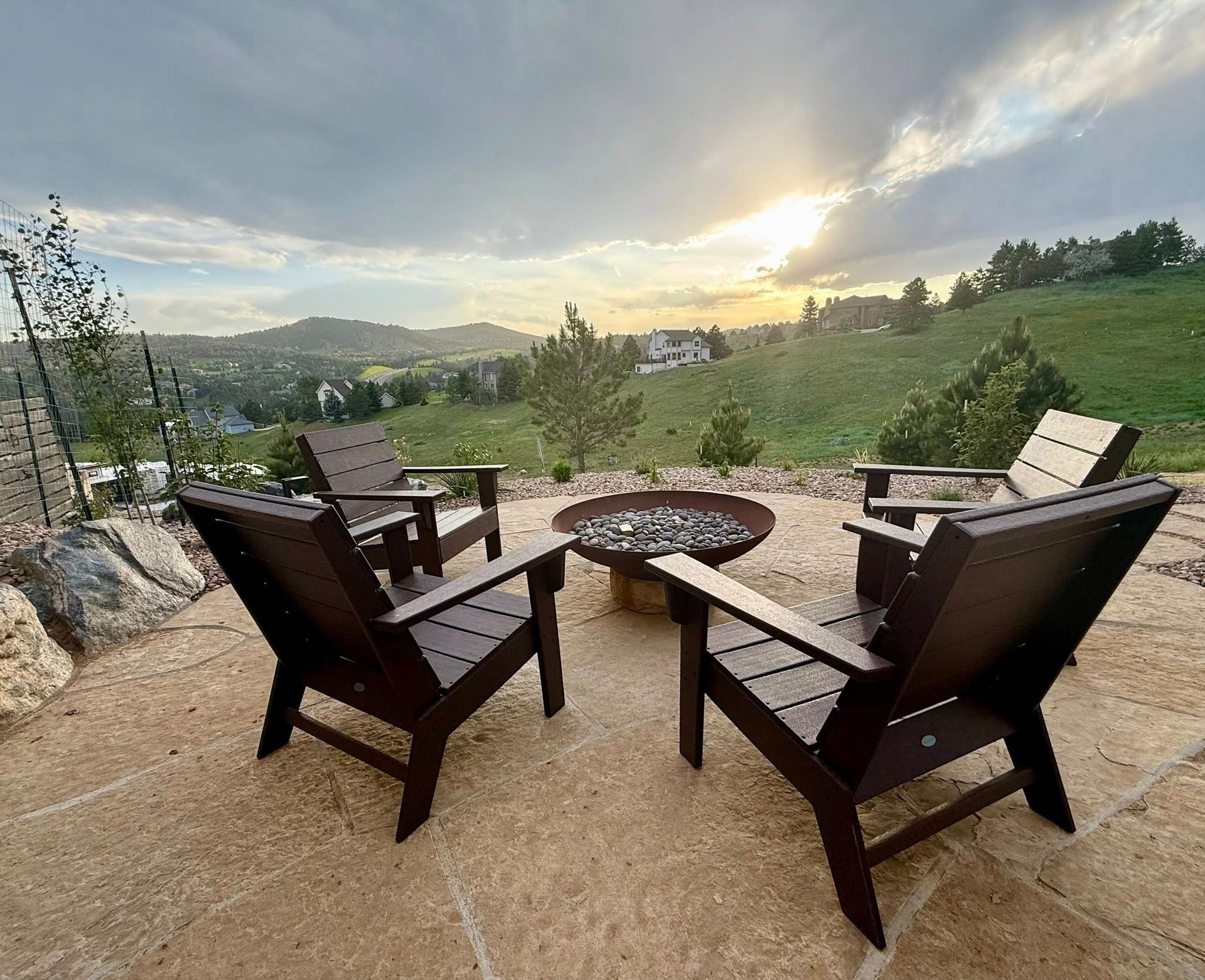 Four dark Adirondack chairs surround a fire pit on a stone patio overlooking a hilly landscape at sunset.