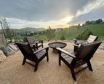 Four wooden chairs surround a fire pit on a stone patio with a scenic mountain sunset.