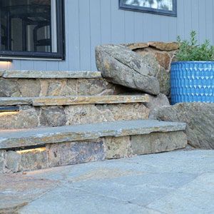 Stone steps leading to a house with a large boulder and blue planter to the side.