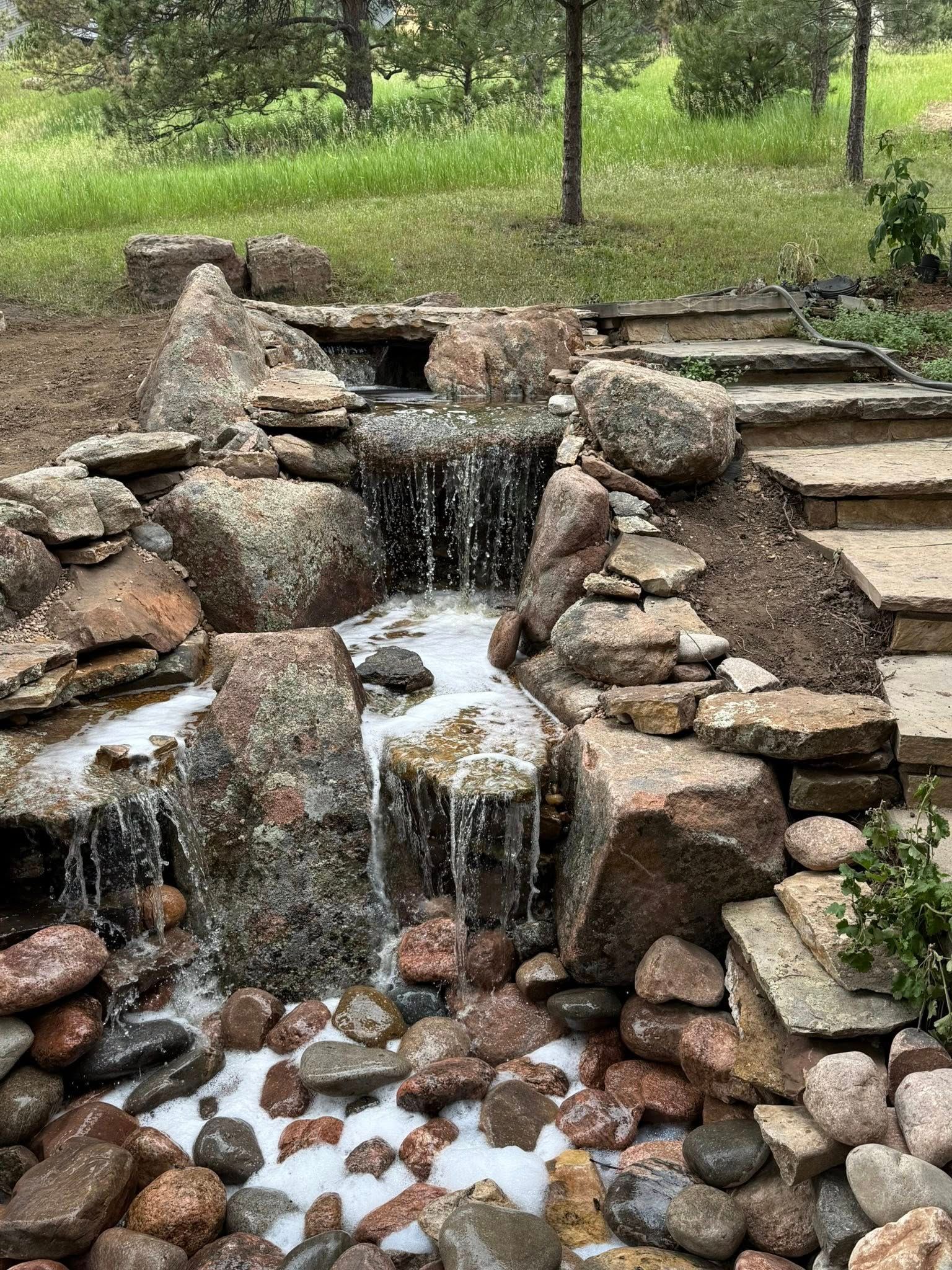Waterfall cascading over rocks into a pool filled with stones. Stone steps ascend to the right, green grass in background.