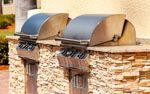 Two stainless steel outdoor grills with raised lids on a stone structure.