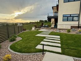 A lush green lawn with stone pavers leads to a modern house. A wooden fence lines the yard with a sunset in the background.