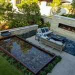 Outdoor patio with water feature, seating, and fireplace. Beige and green tones, sunny setting.