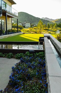 Landscaped yard with a water feature, stone walls, blue flowers, and green lawn. Mountains in the background.